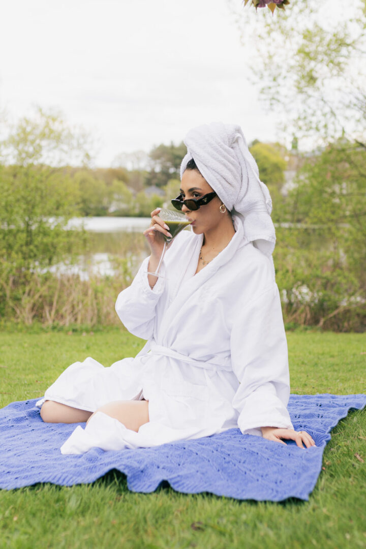 A woman in white robe sitting on blue blanket near body of water.